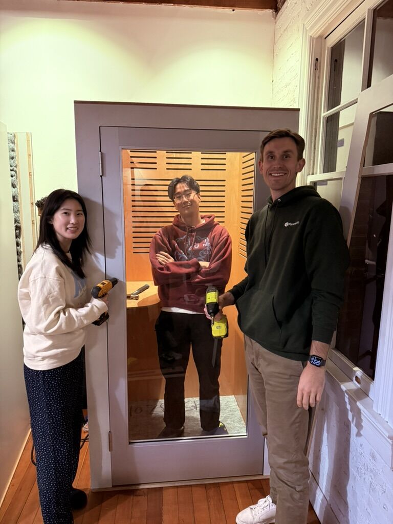 Noah Plewa, on the right, holding a cordless drill beside two people and a glass-fronted booth