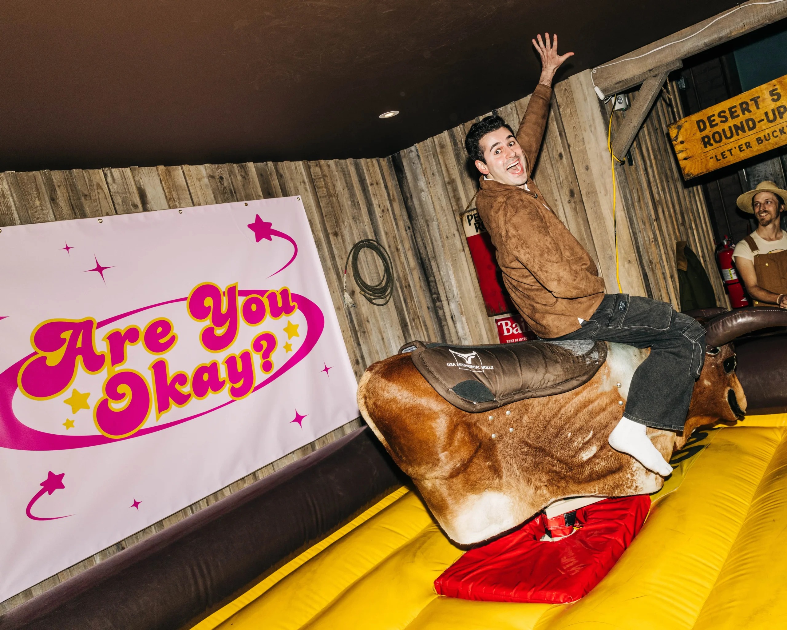 A guest rides a mechanical bull beside an Are You Okay? sign at a NowThis event