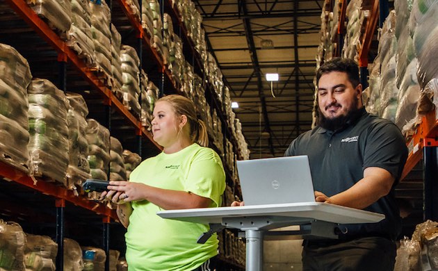 Two warehouse workers use a handheld scanner and laptop beside pallet racks