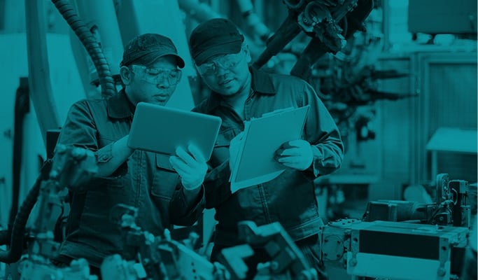 Two factory workers review a tablet beside production equipment