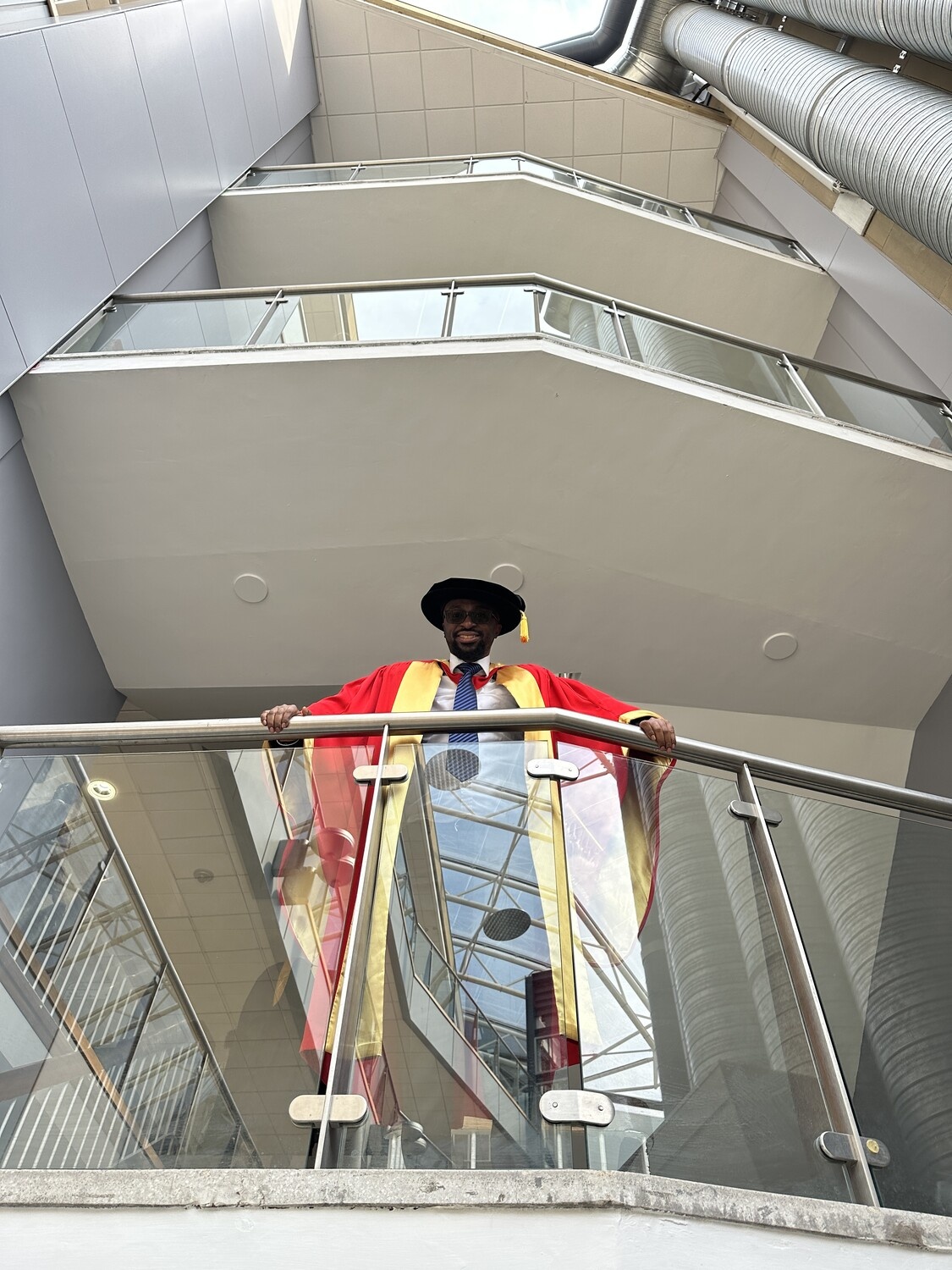 Odiong Akpan in red and gold academic robes, standing on a balcony beneath several higher floors