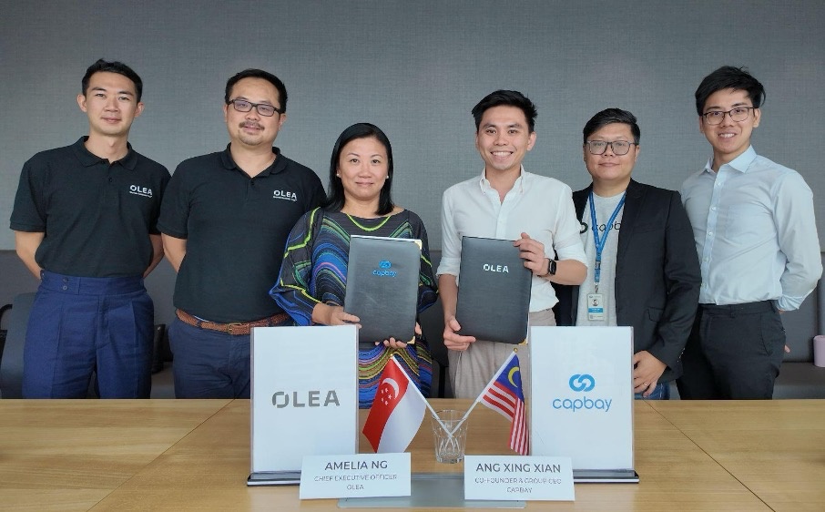 Olea and CapBay representatives celebrate their partnership, with Amelia Ng and Ang Xing Xian holding signed folders