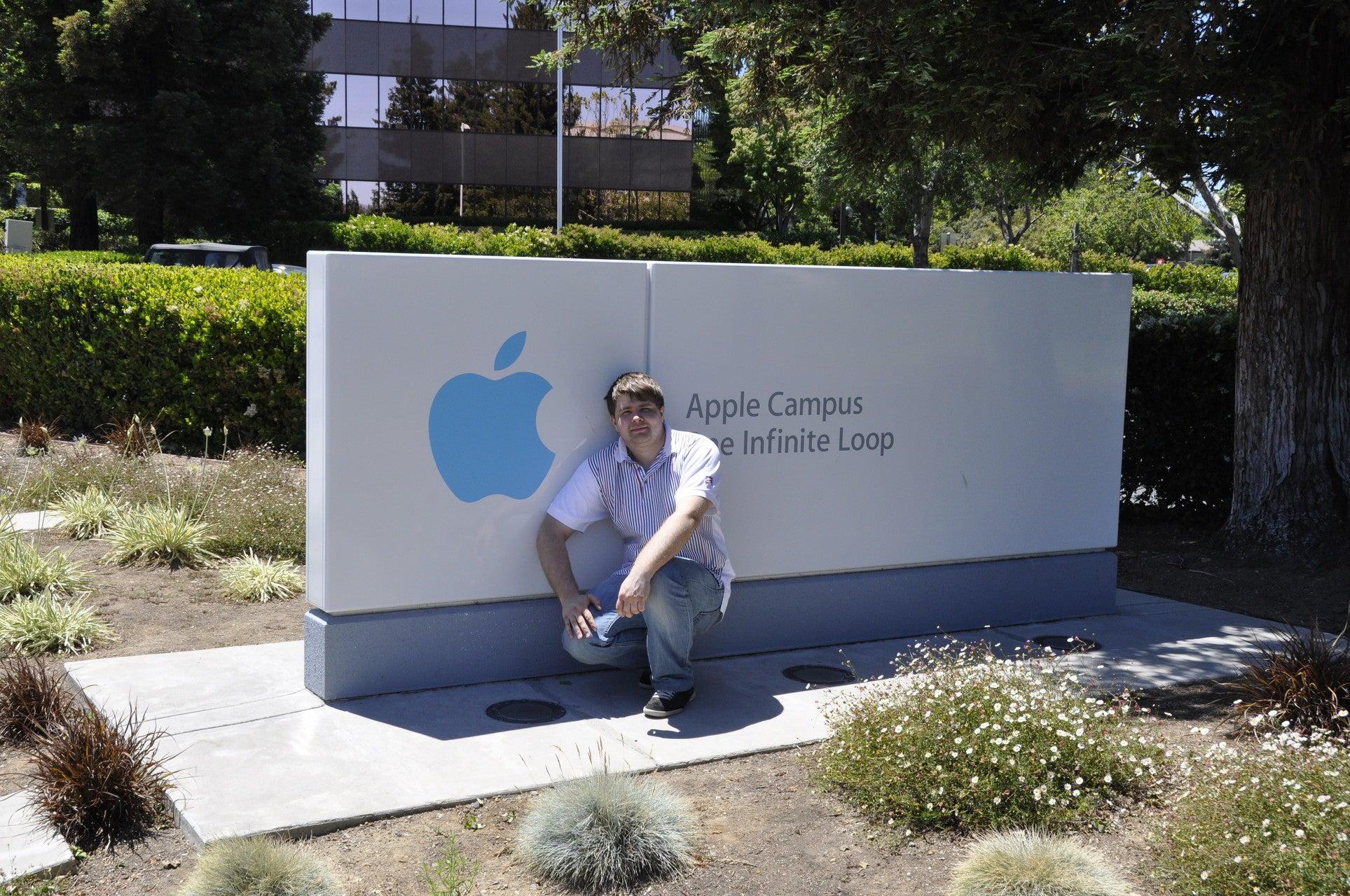 A younger Oleksandr Kosovan crouching beside the Apple Campus sign at One Infinite Loop