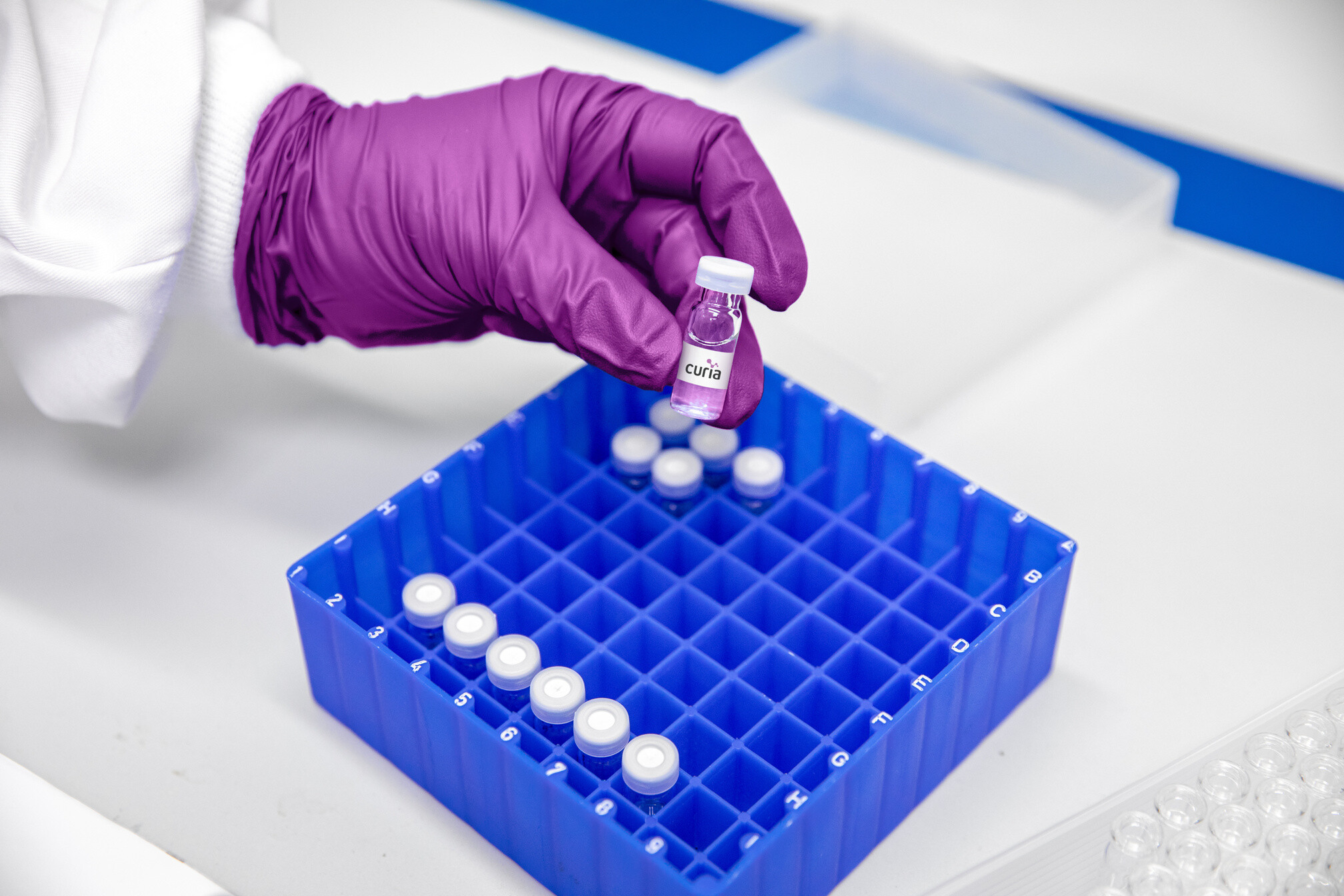 A gloved Curia laboratory worker holds a small vial above a blue sample rack