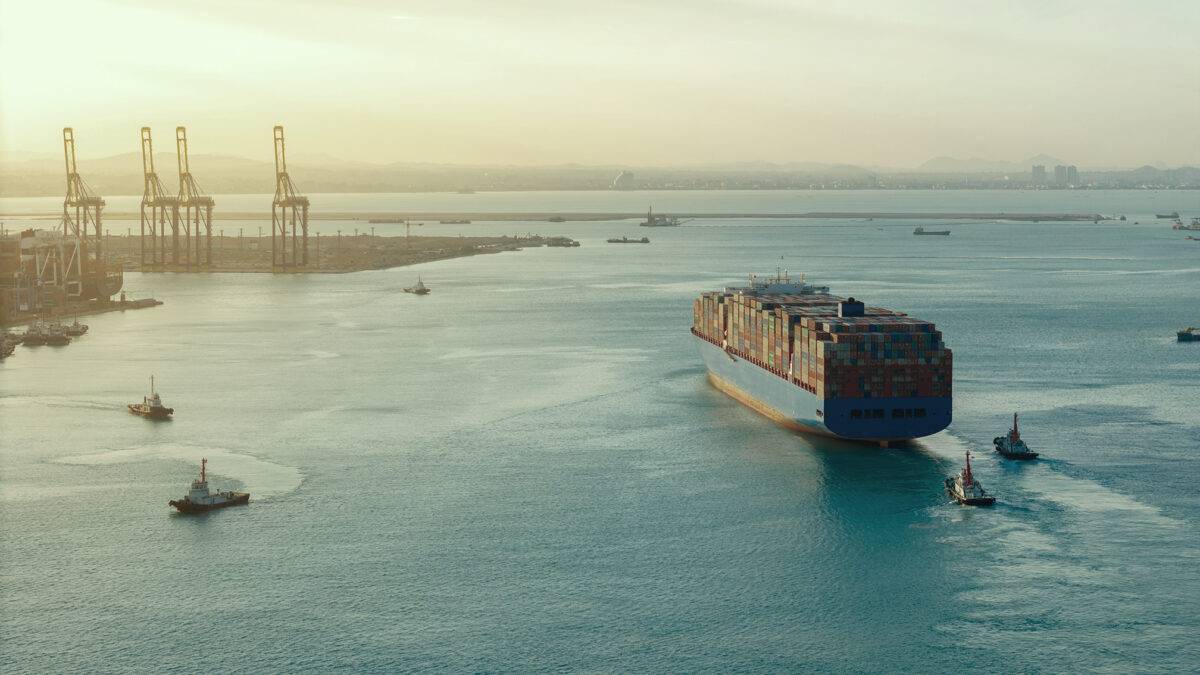 A container ship moves through a port accompanied by tugboats