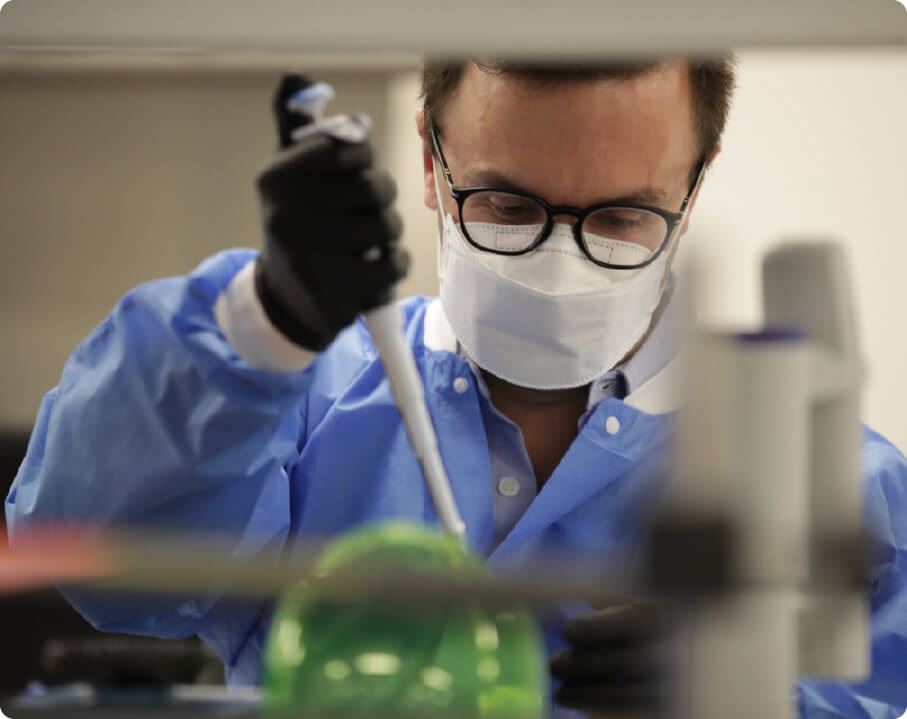 A scientist wearing glasses, a mask and gloves pipettes liquid at a laboratory bench
