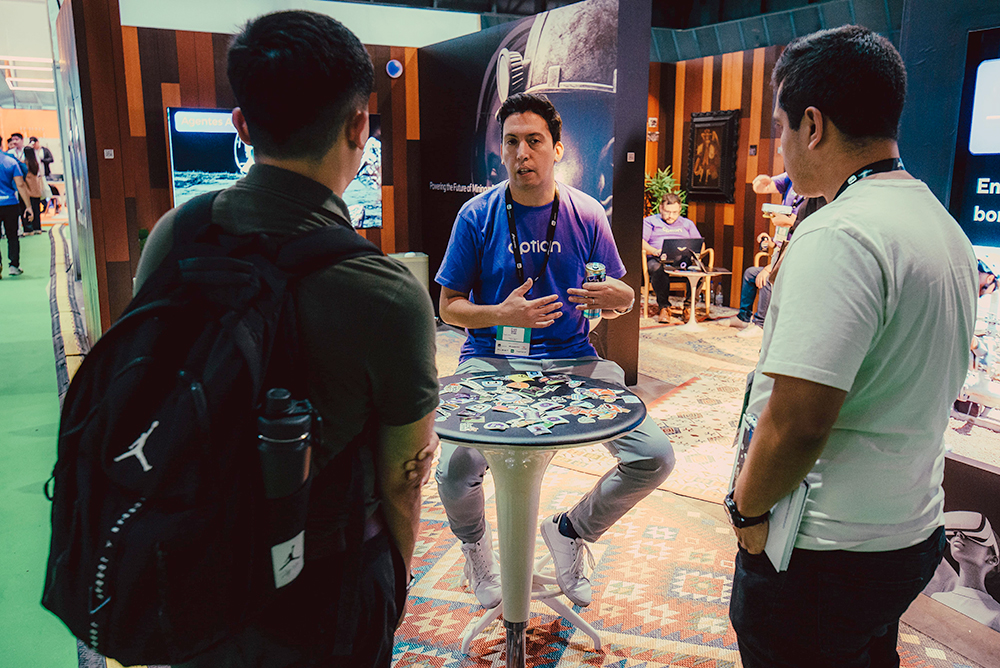 An Option representative speaking with visitors around a sticker-covered table at Expomin 2025