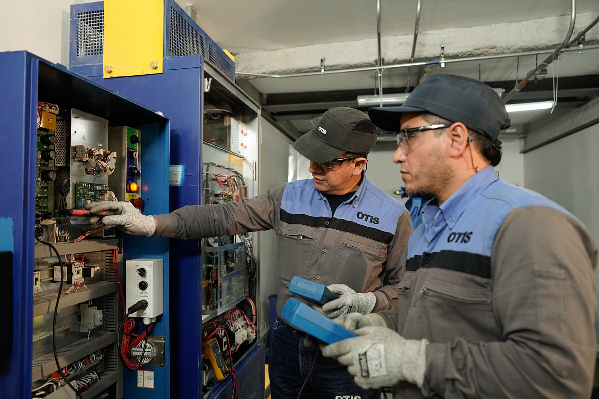 Two Otis technicians inspect electrical components and use diagnostic instruments in an elevator machine room
