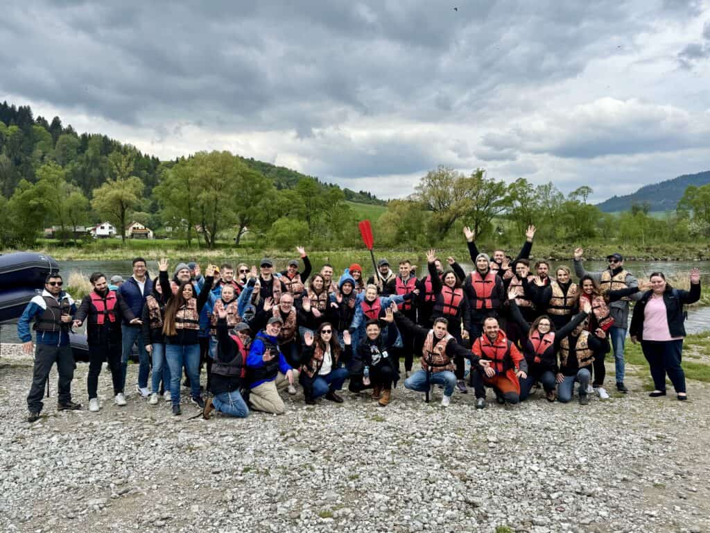 The Oversee team gathered by a river on an offsite, wearing life jackets and waving