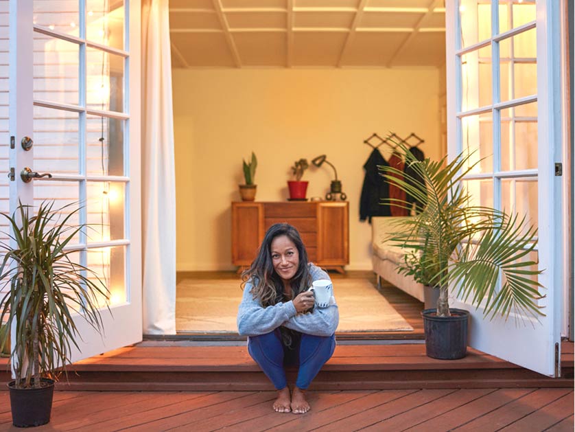 A woman holding a mug sits barefoot on the steps of an open, warmly lit home