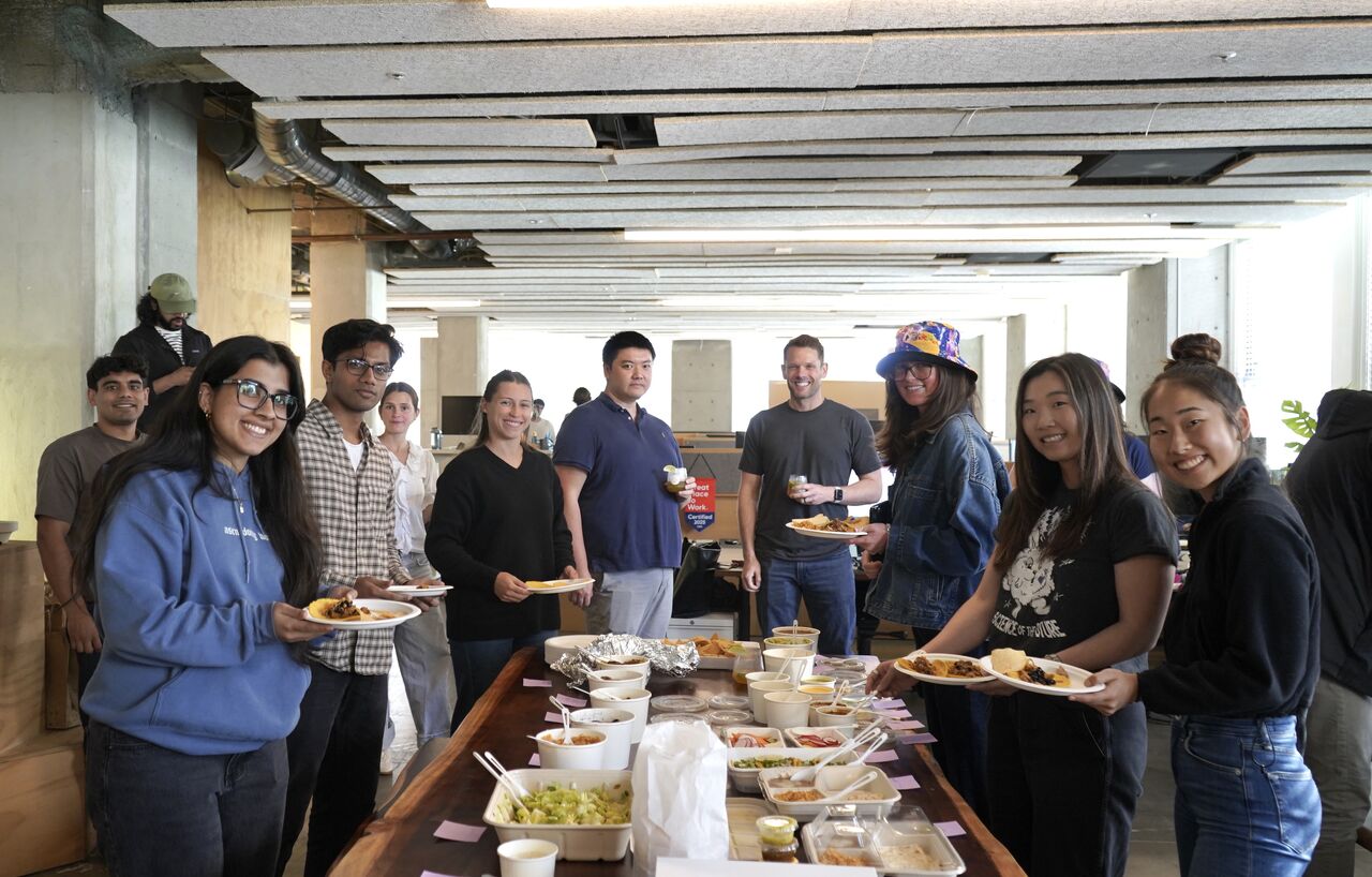 The Pallet team gathered around a long table of food in their San Francisco office, smiling.