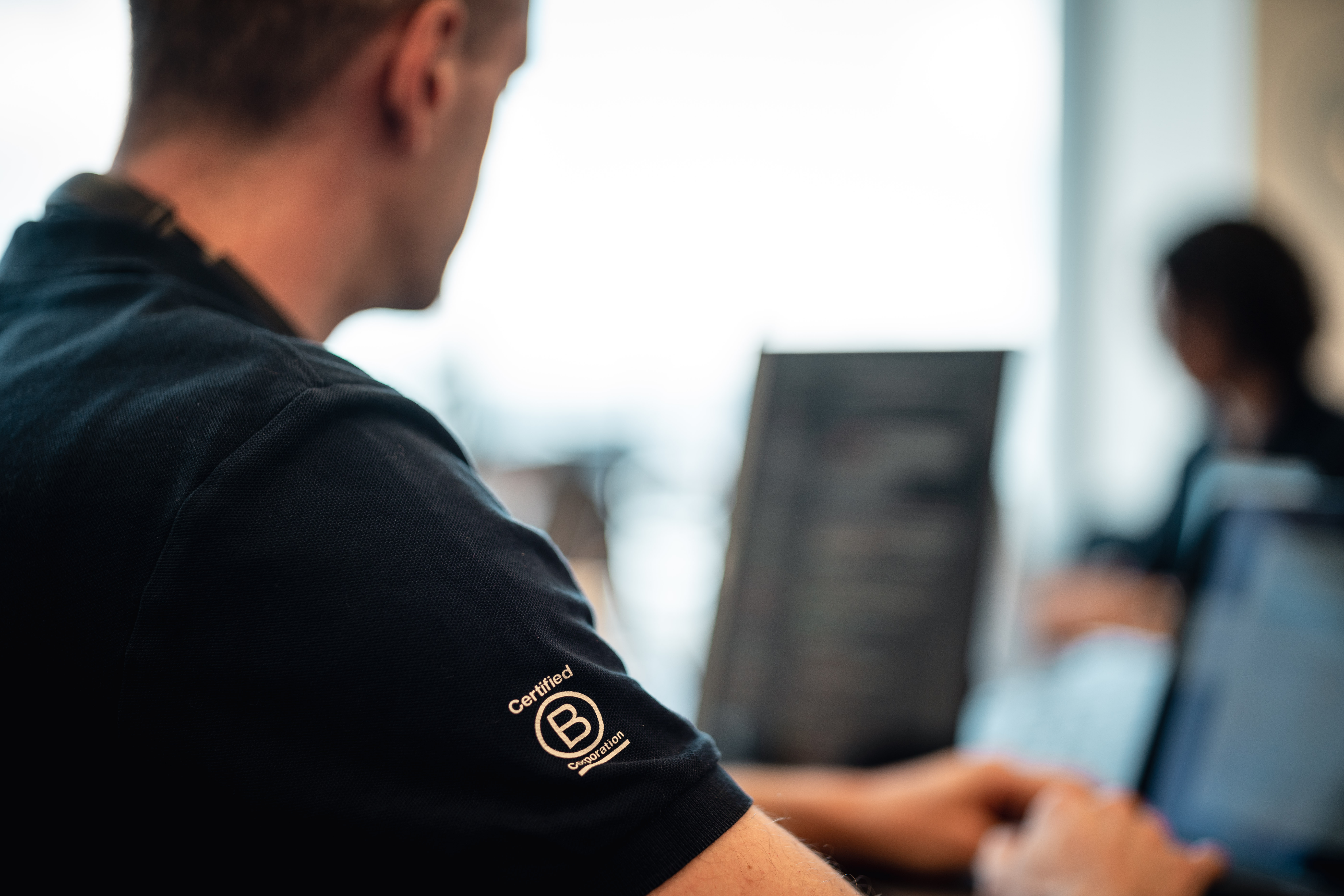 A PALO IT team member at a laptop wearing a shirt with a B Corp insignia