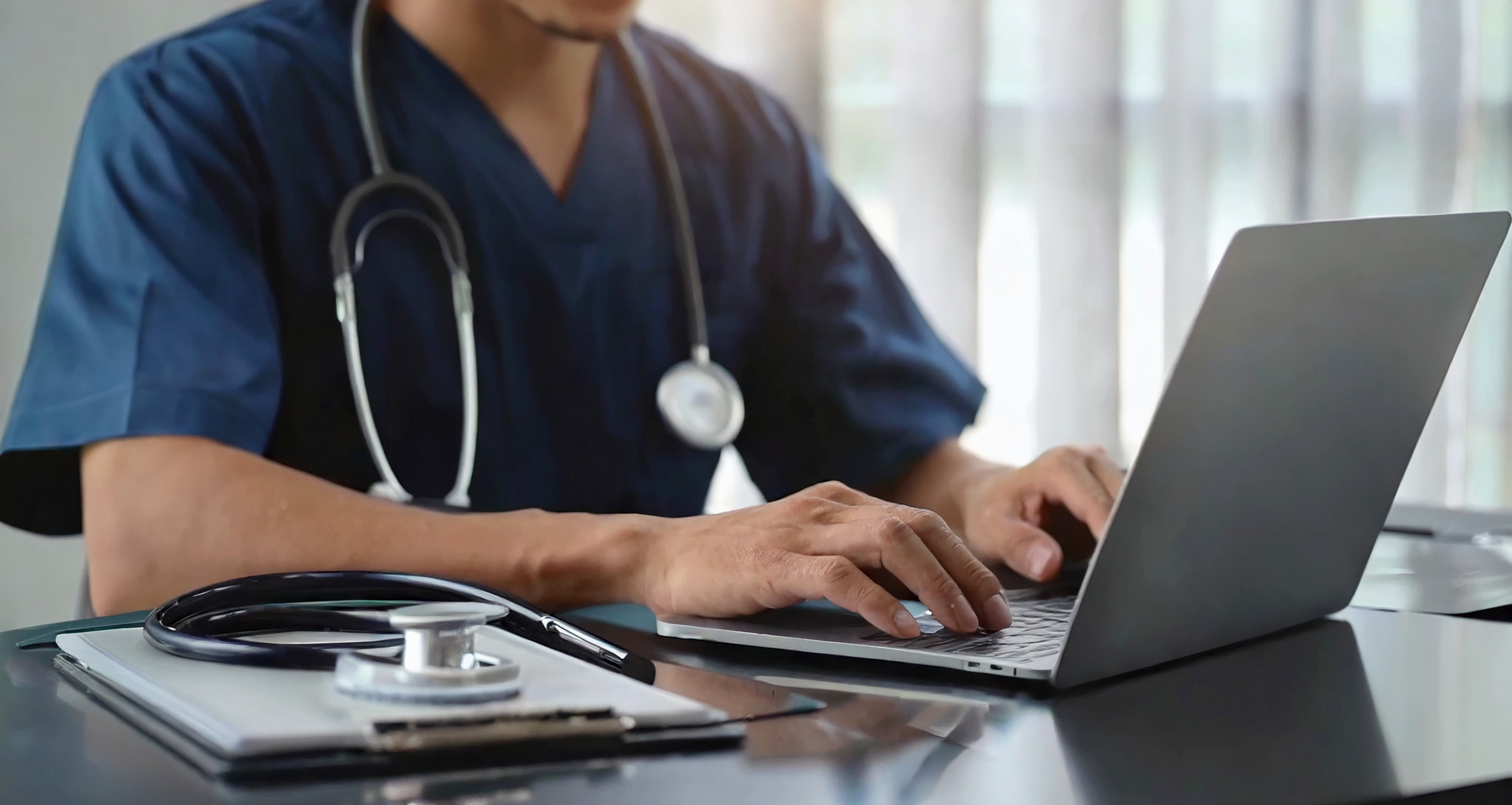 A clinician in scrubs working on a laptop with a stethoscope nearby