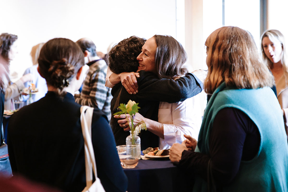 Two women embracing at a lively Parsons community gathering while other guests talk around them