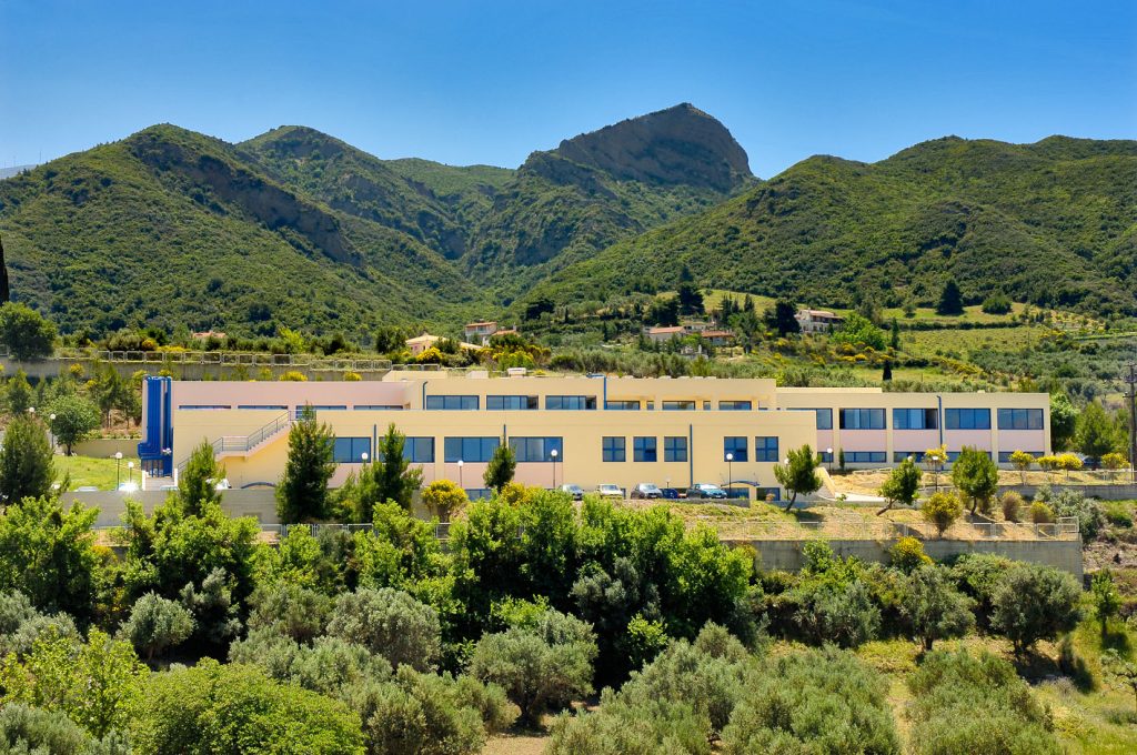 The pale-yellow Patras Science Park buildings, with parked cars and green hills behind them