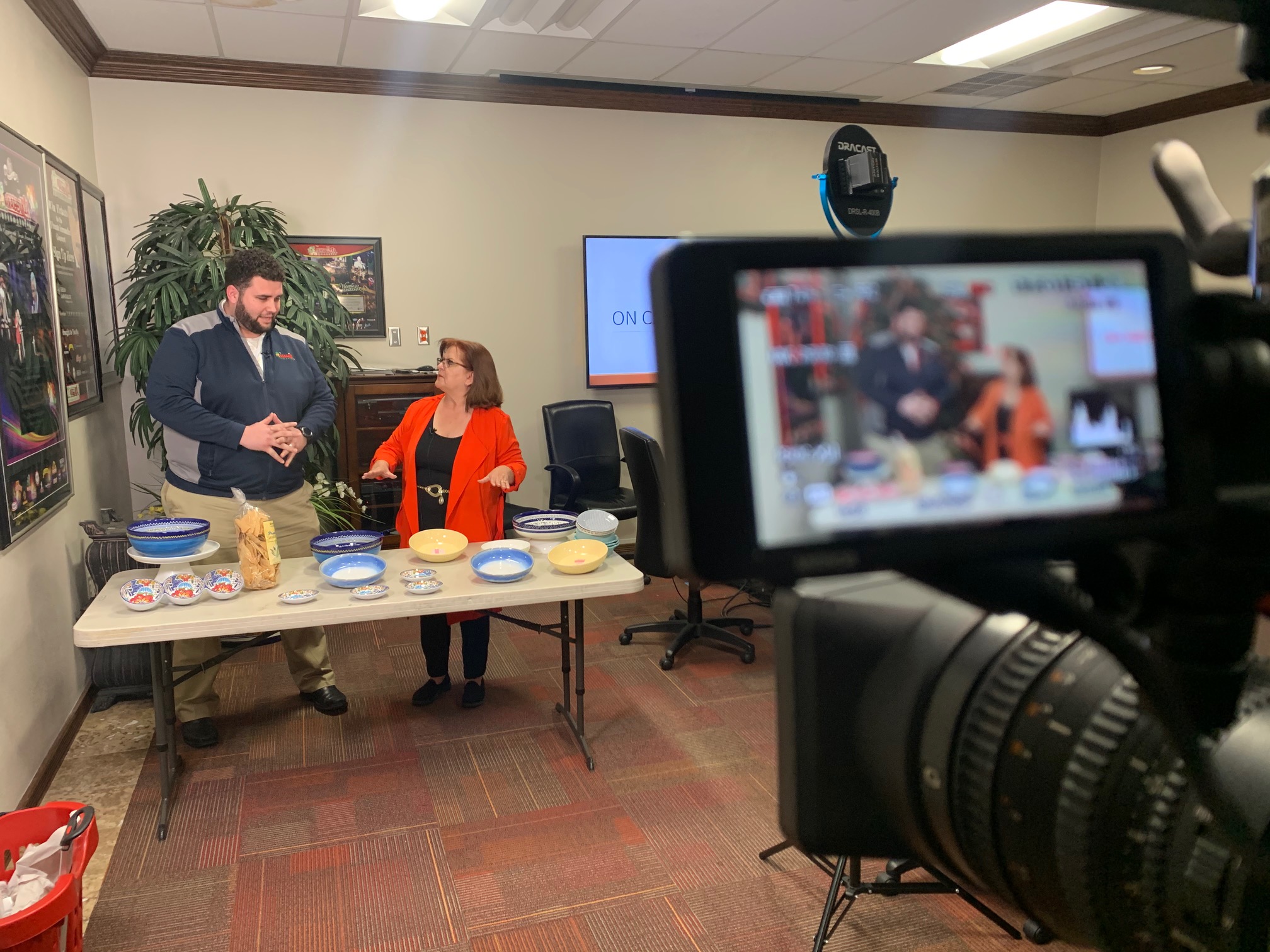 Patricia Pérez in an orange jacket during a filmed demonstration, seen beside a table and through a camera monitor