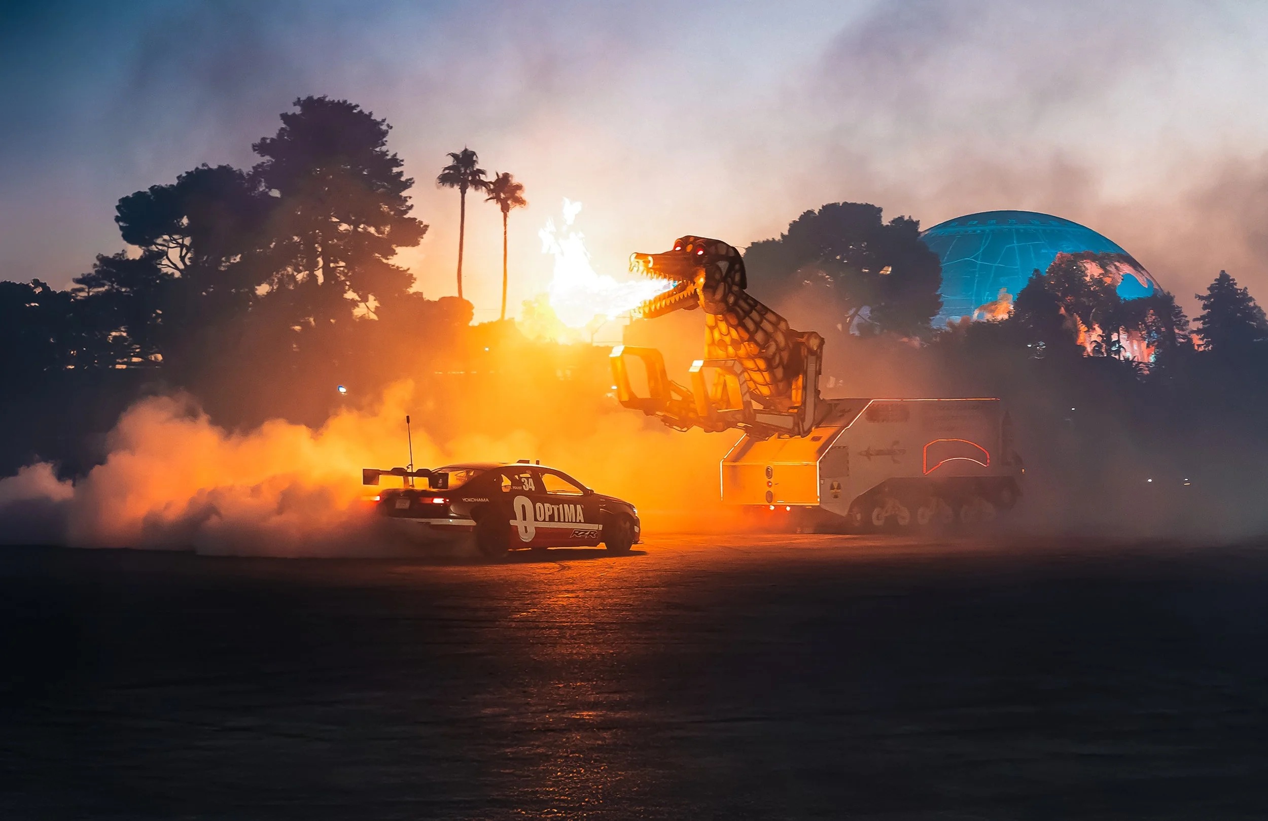 An OPTIMA-branded drift car alongside a mechanical fire-breathing dinosaur at an event