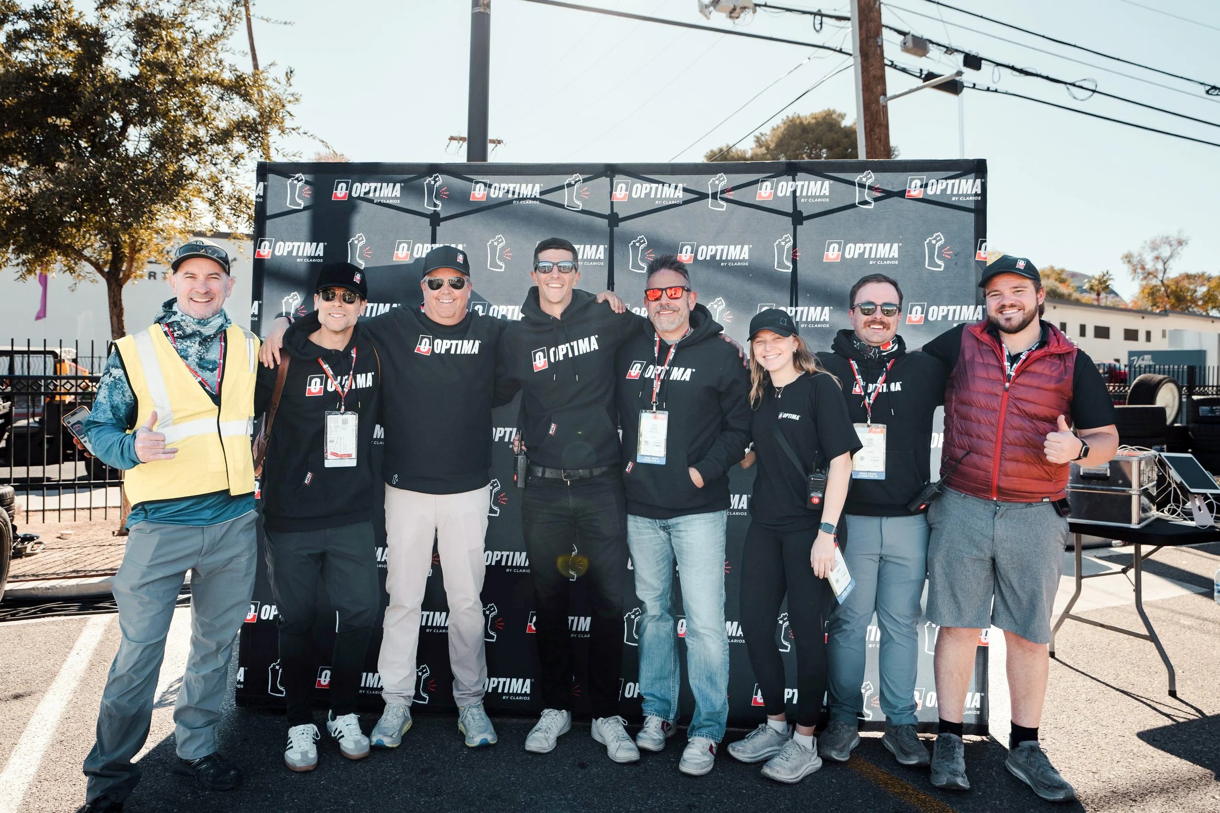 A group in OPTIMA event apparel posing together in front of a branded backdrop
