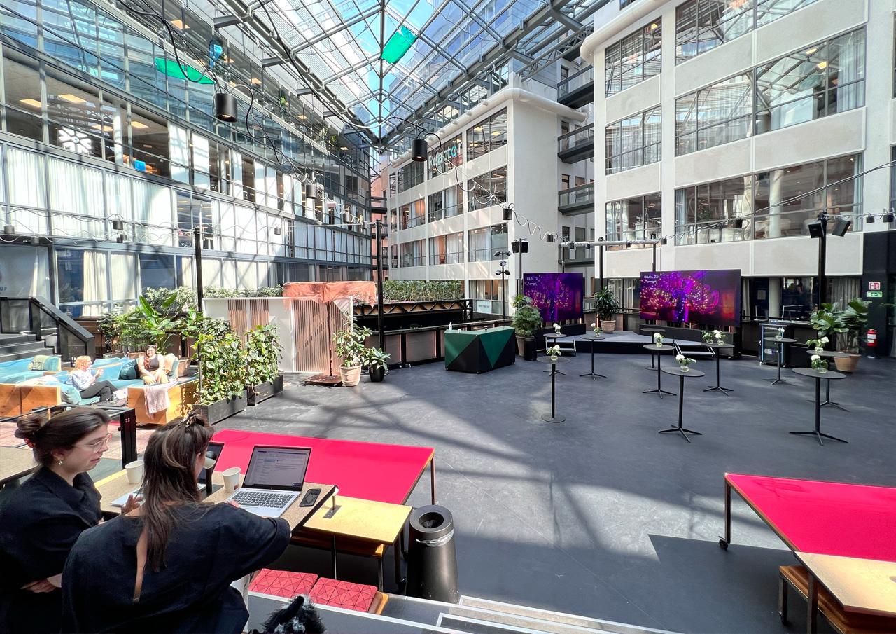 Shared tables, seating and an event stage beneath Epicenter Stockholm’s glass-roofed atrium