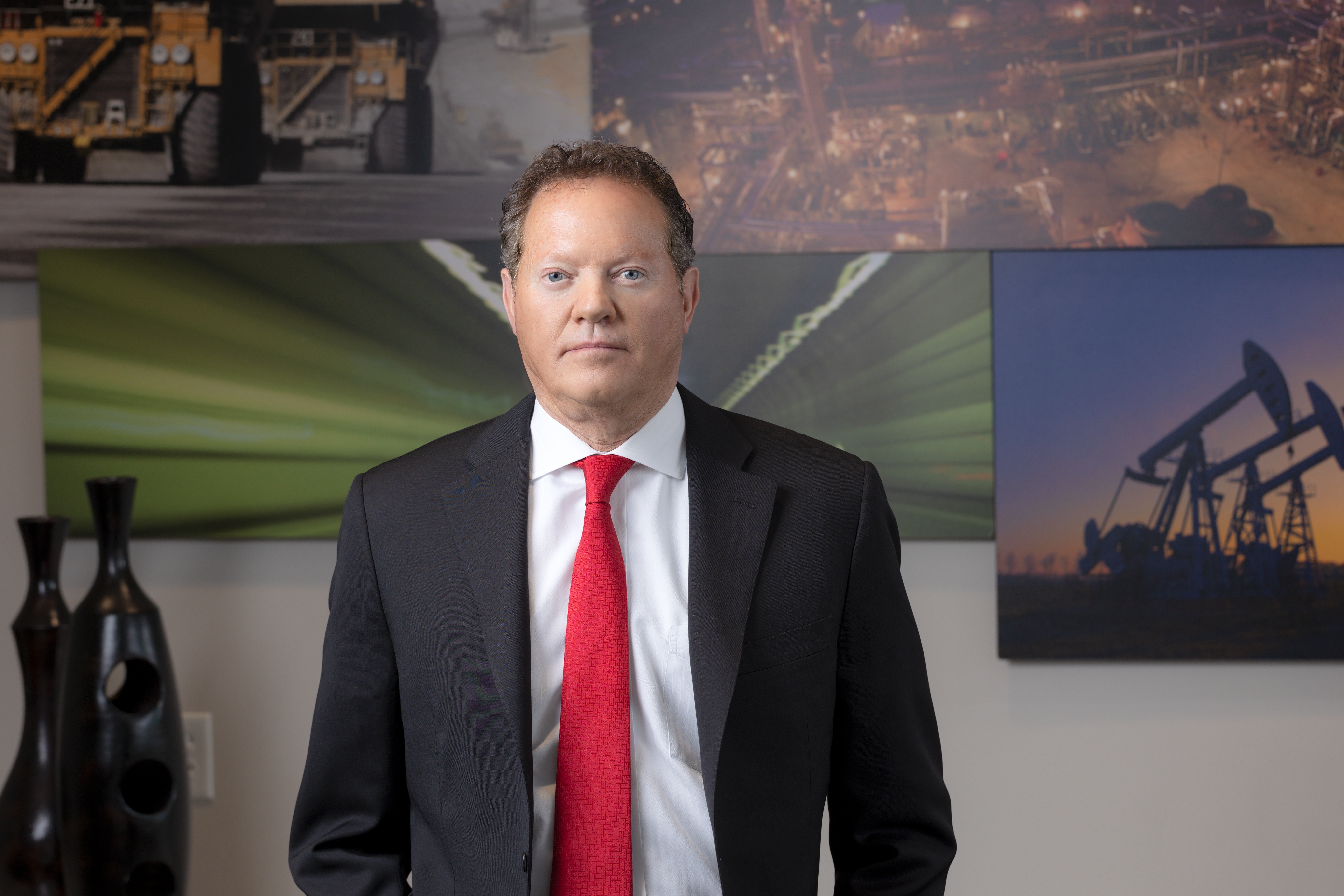 Patrick Williams in a dark suit and red tie, standing before photographs of mining equipment and energy operations