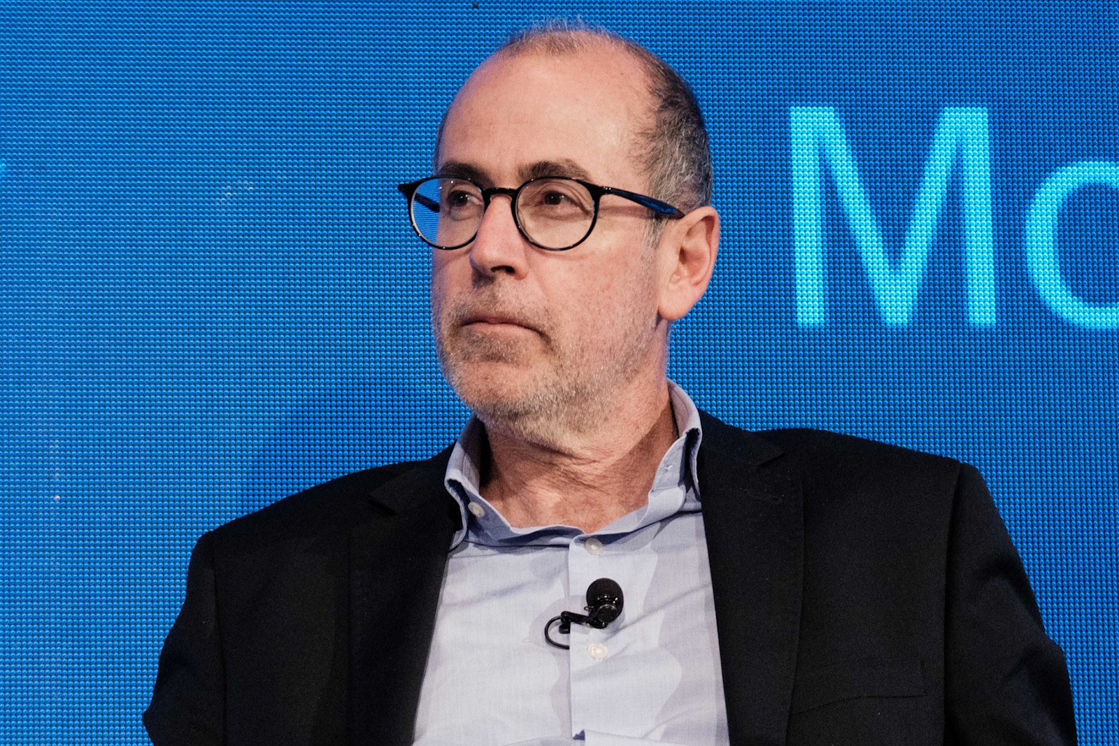Paul Bassat seated at a business event, wearing glasses and a lapel microphone against a blue backdrop