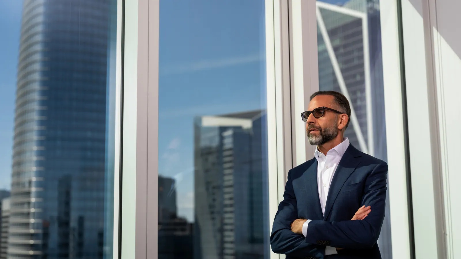 Paul Bresge in a dark suit, standing by a window overlooking a city