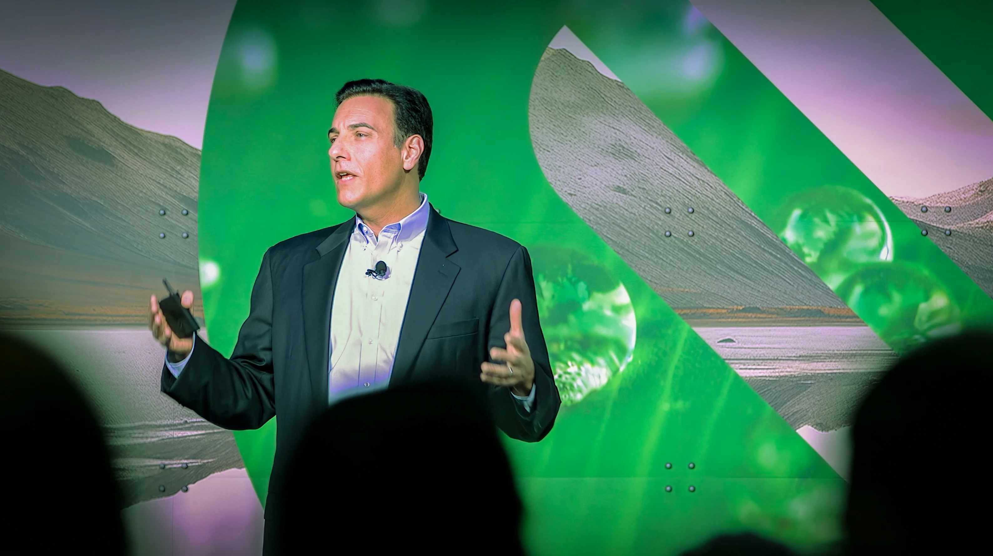 Paul Marushka speaking onstage with a presentation remote against a green event backdrop