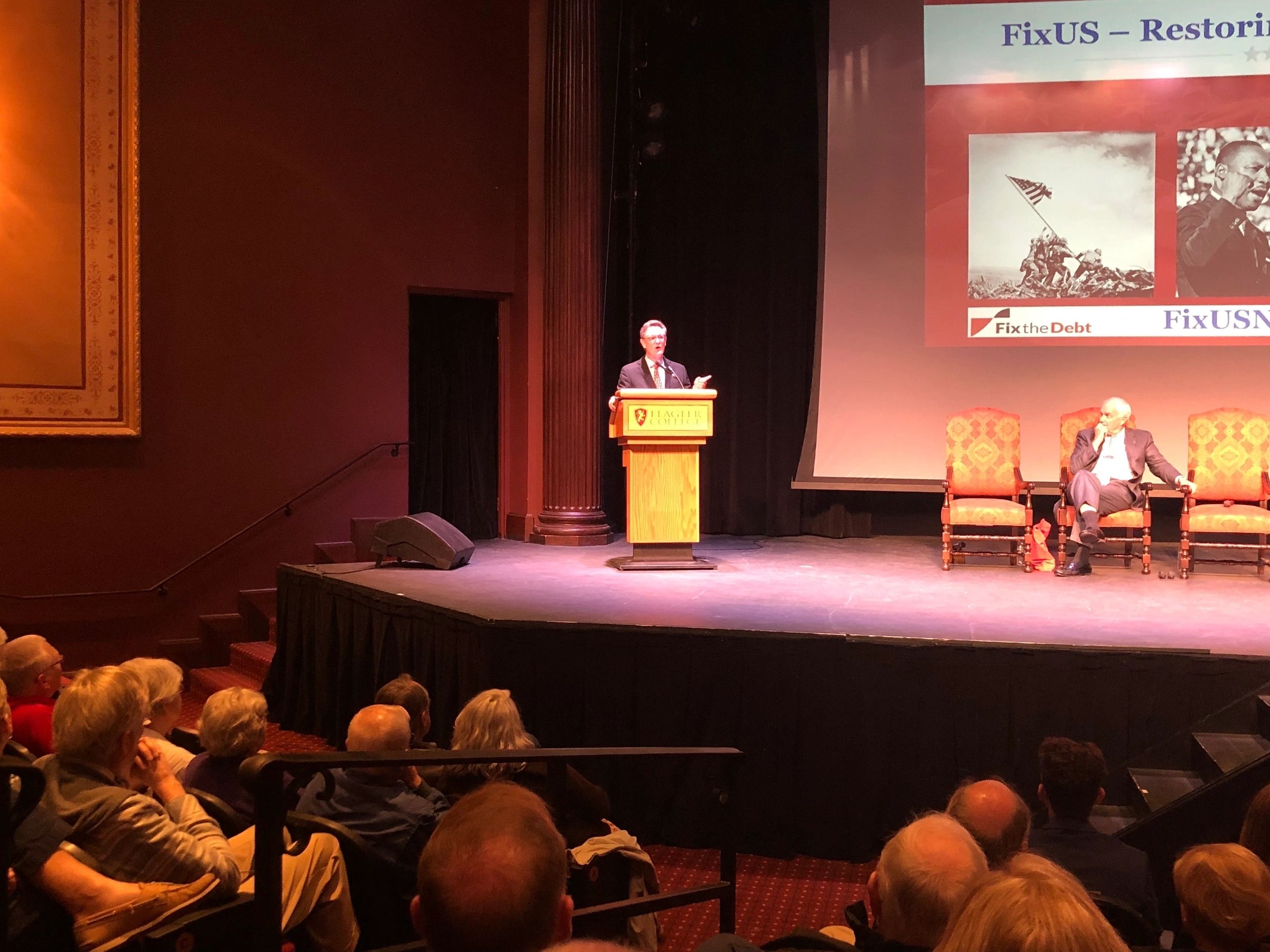 Paul Stebbins at a lectern addressing an audience at Flagler College, with a FixUS presentation projected behind him.