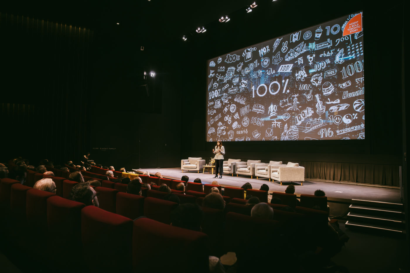 Paula Port presents Destination Toronto's 100% campaign on a theatre stage before a seated audience