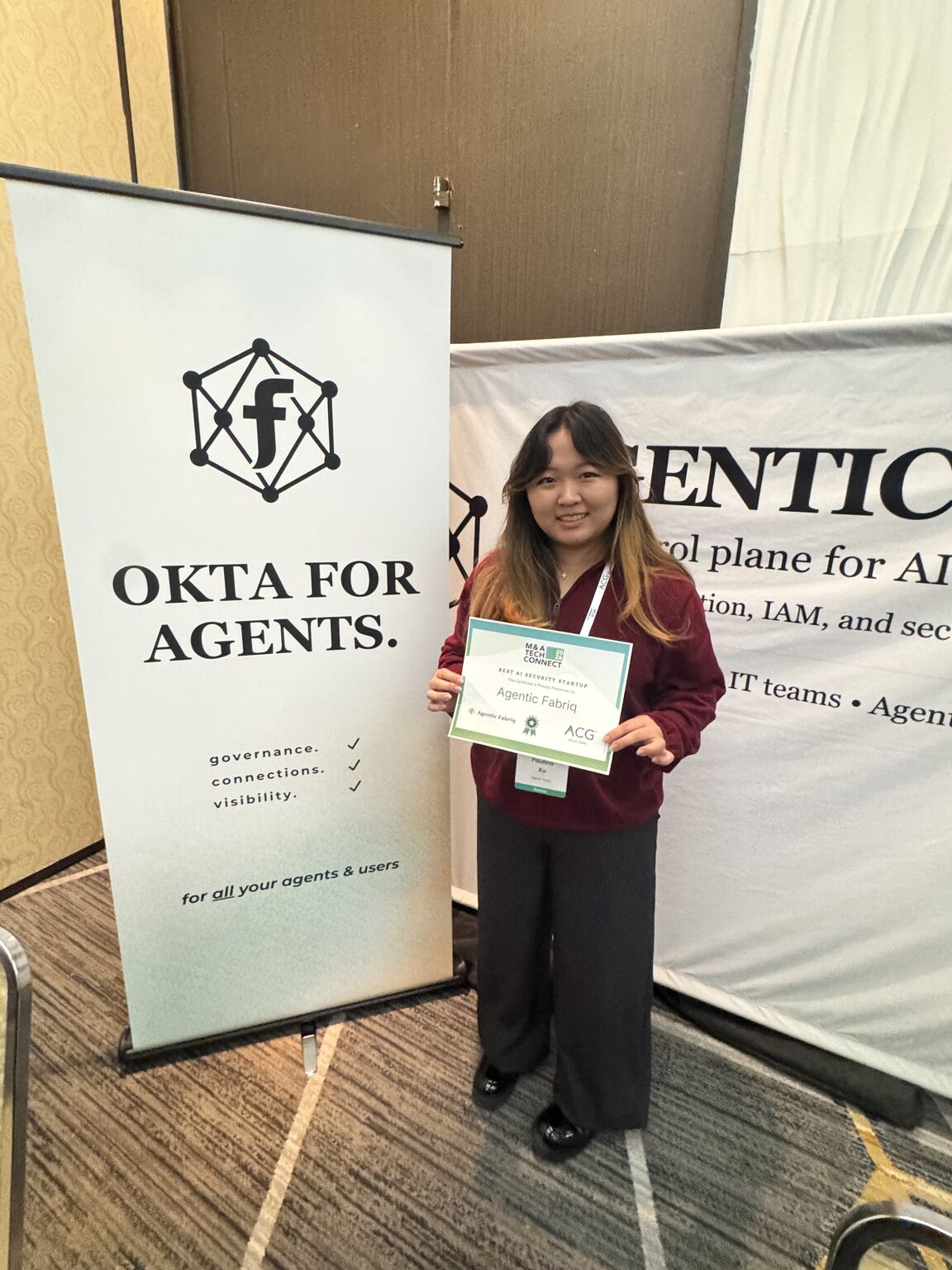 Paulina Xu stands beside the Agentic Fabriq booth at a 2026 conference holding an exhibitor sign