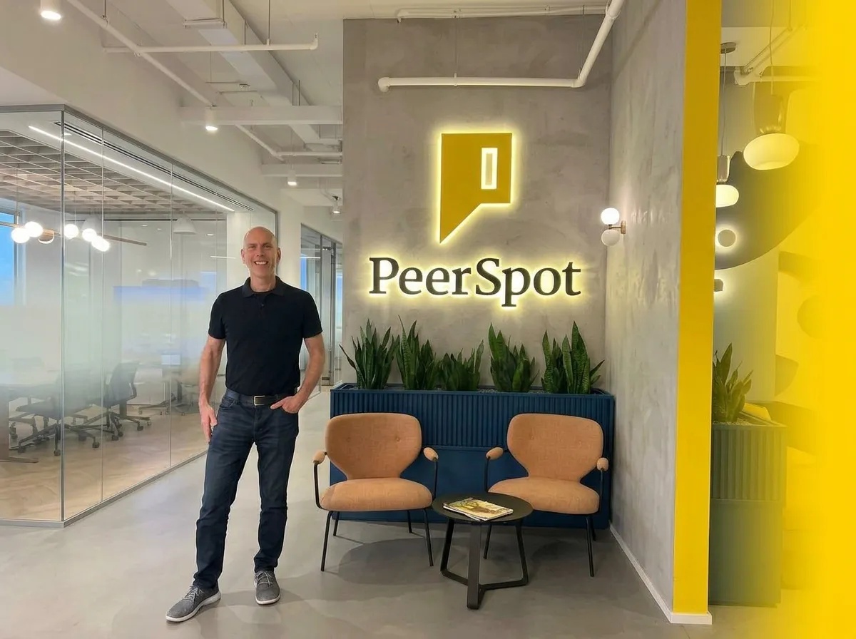 PeerSpot co-founder Russell Rothstein standing in an office beside two chairs and the company sign