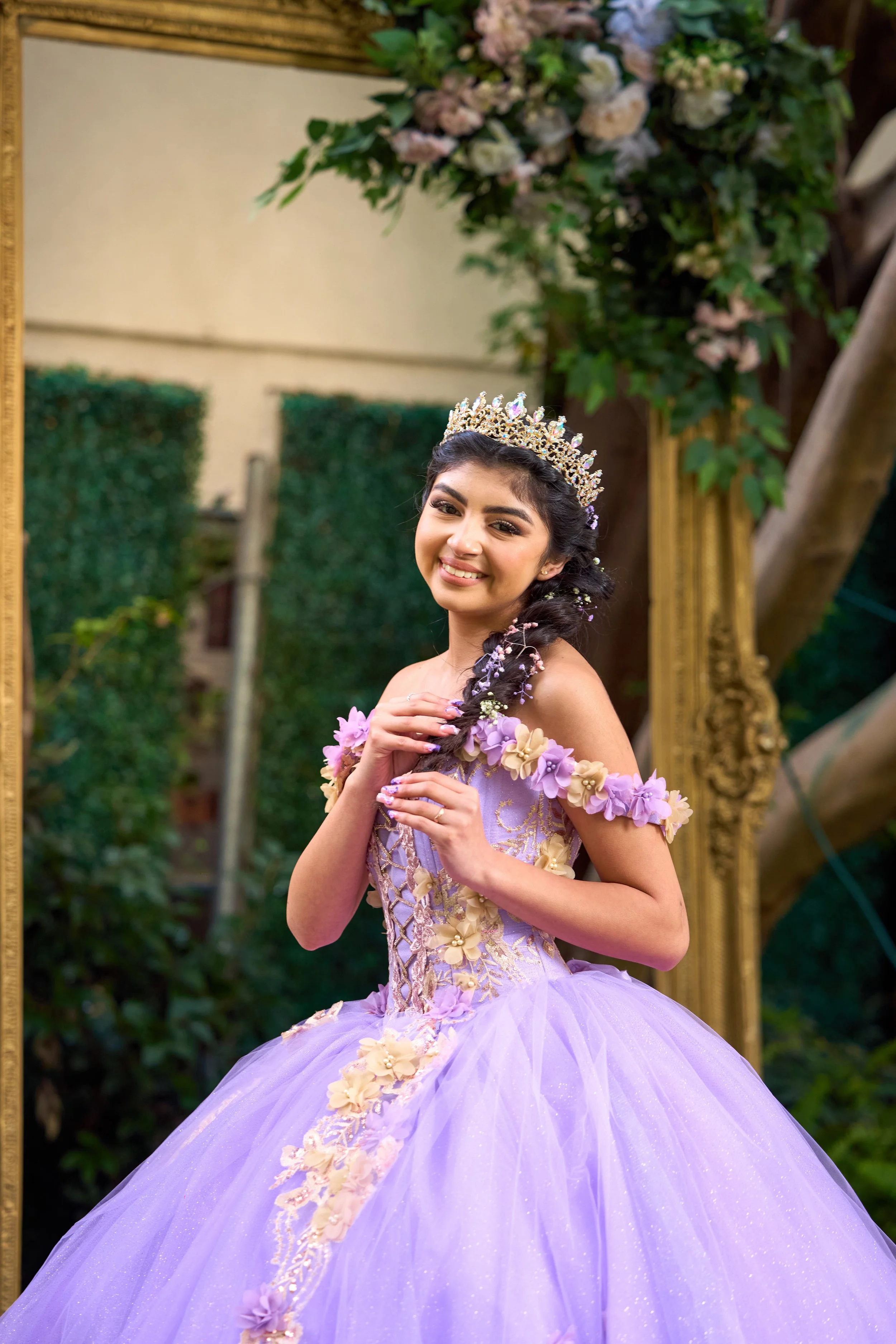 A celebrant in a lavender gown with a flower-threaded braid, photographed against greenery and a gold frame