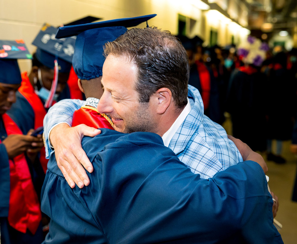 Pete Kadens embraces a graduate in a blue cap and gown.