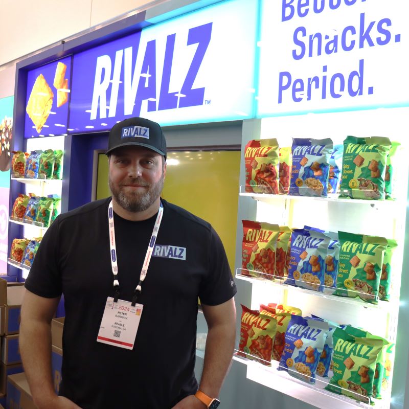 Peter Barrick wearing Rivalz clothing at the company's Expo West booth, with colorful snack bags displayed behind him