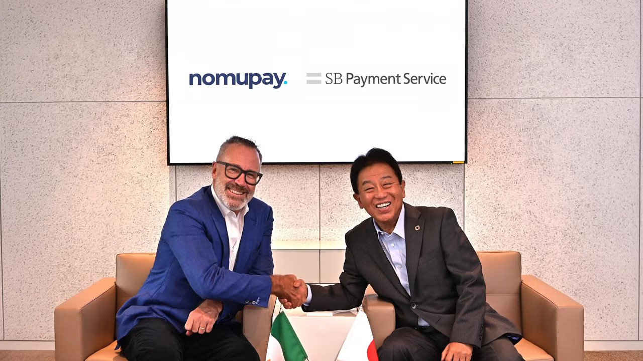 Peter Burridge, left, and Jun Shimba shake hands beneath Nomupay and SB Payment Service signage.