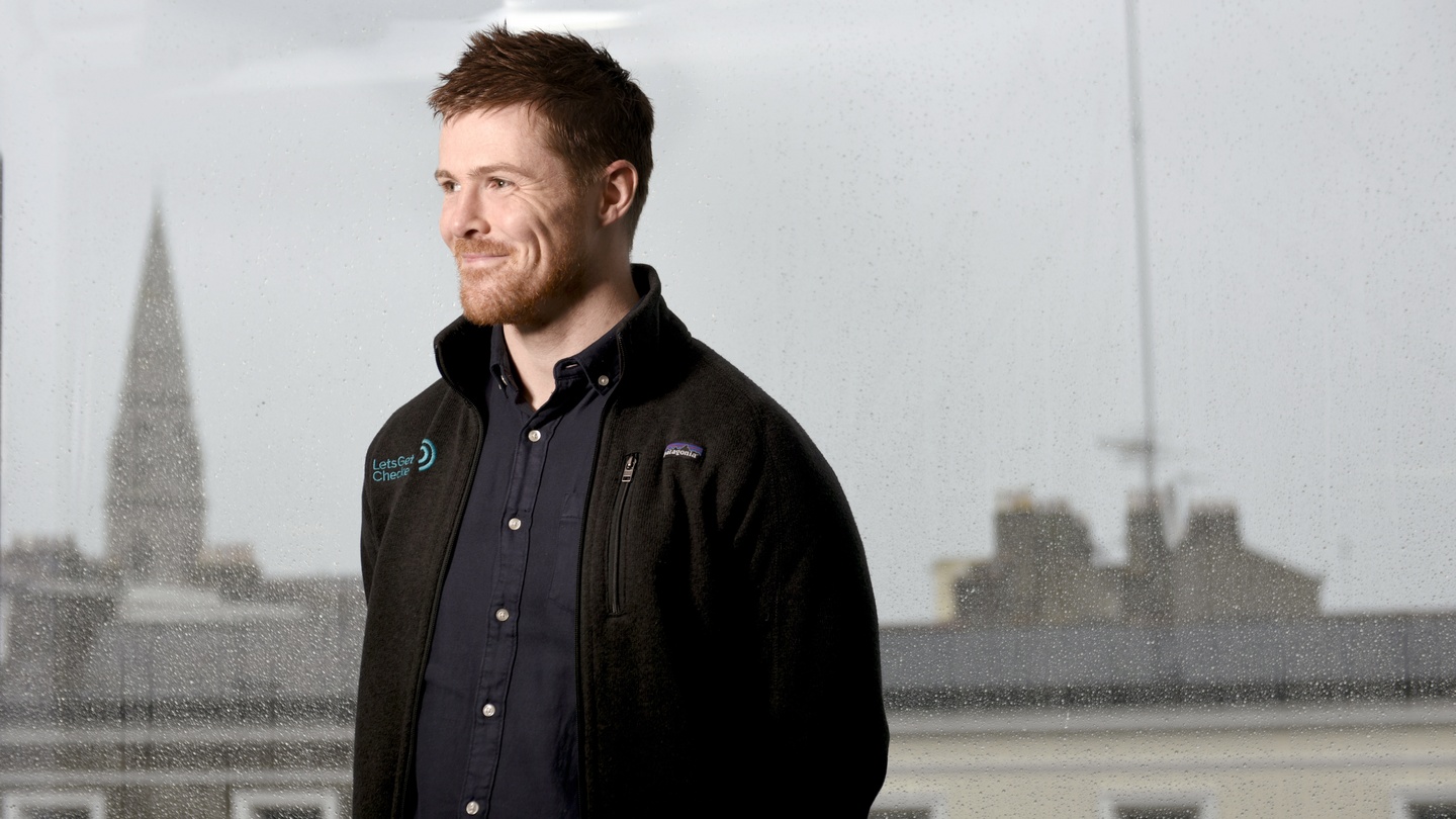 Peter Foley beside a rain-speckled office window, wearing a LetsGetChecked jacket