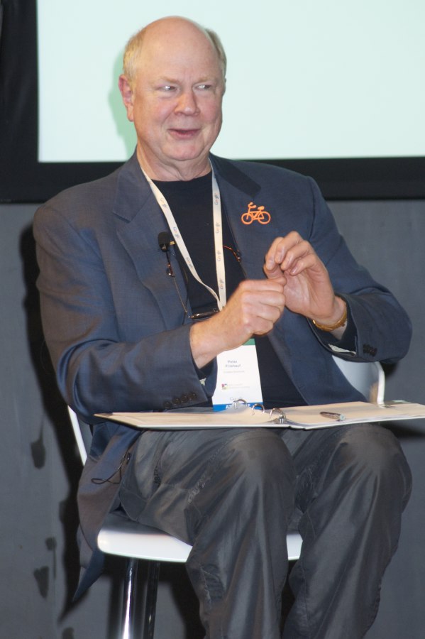 Peter Frishauf seated with a binder, moderating a discussion, wearing an orange bicycle pin