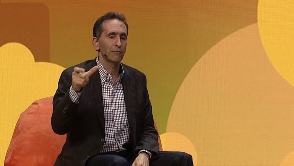 Peter Gassner seated onstage, gesturing during a SaaStr conversation against an orange backdrop.