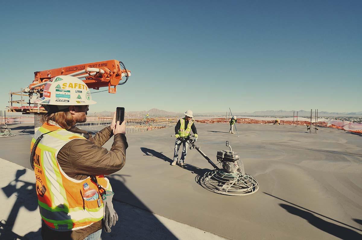 A construction worker records activity on a phone beside a concrete finishing crew