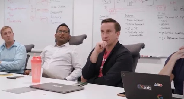 Peter Rex seated at a conference table with colleagues, with planning notes on a whiteboard behind them