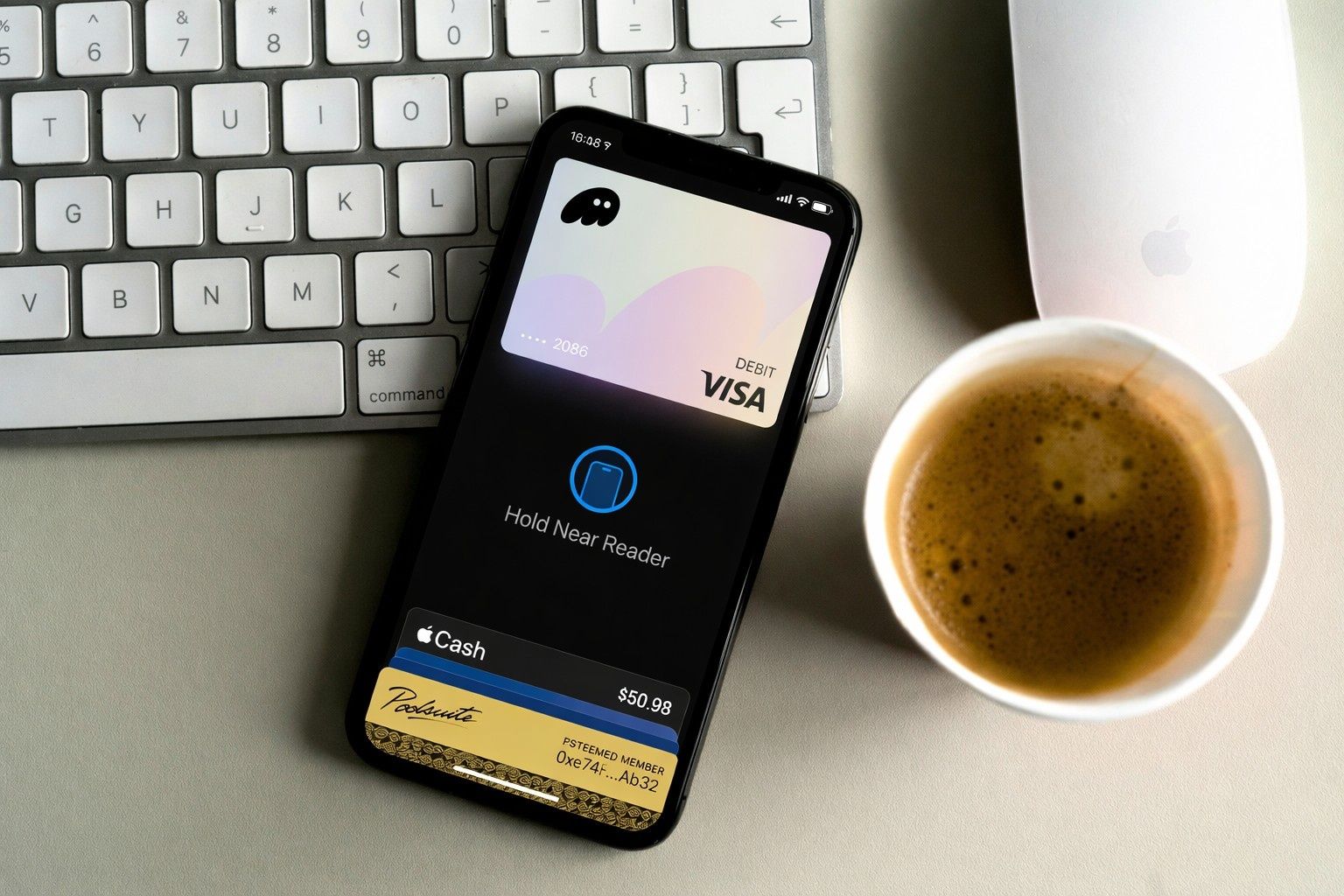 An iPhone showing the Phantom debit card in Apple Pay beside a keyboard, mouse and coffee