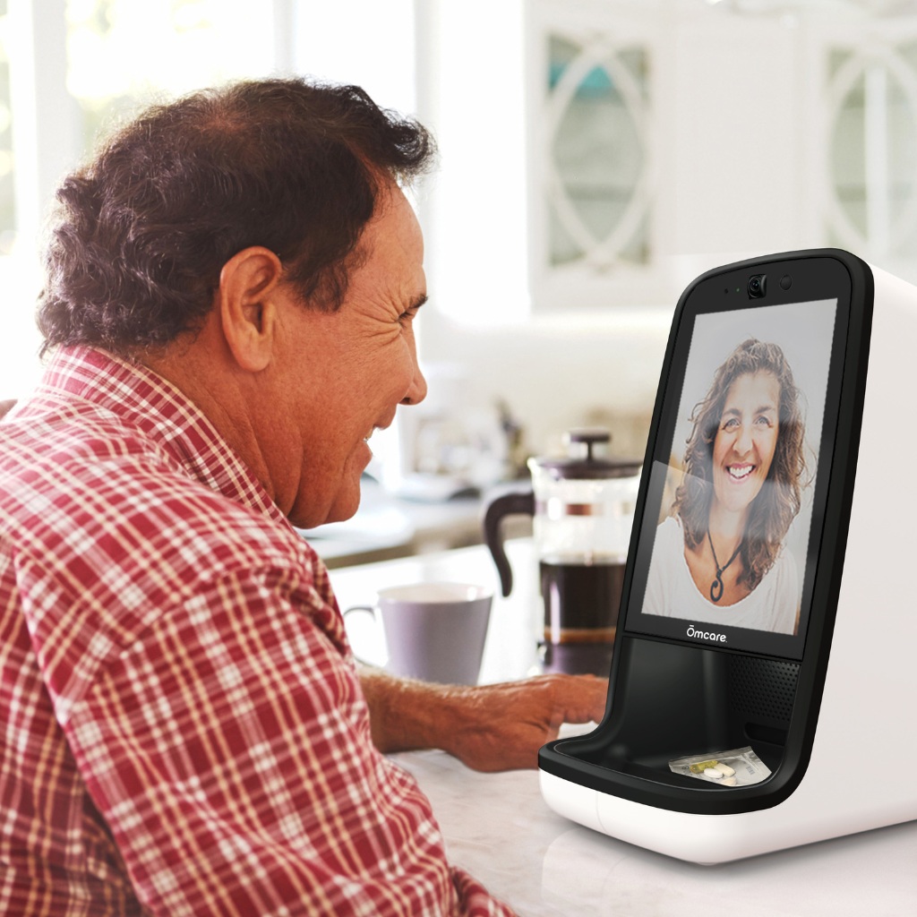 Ōmcare promotional photograph of a man using the Home Health Hub for a video call, with a medication pouch in its tray