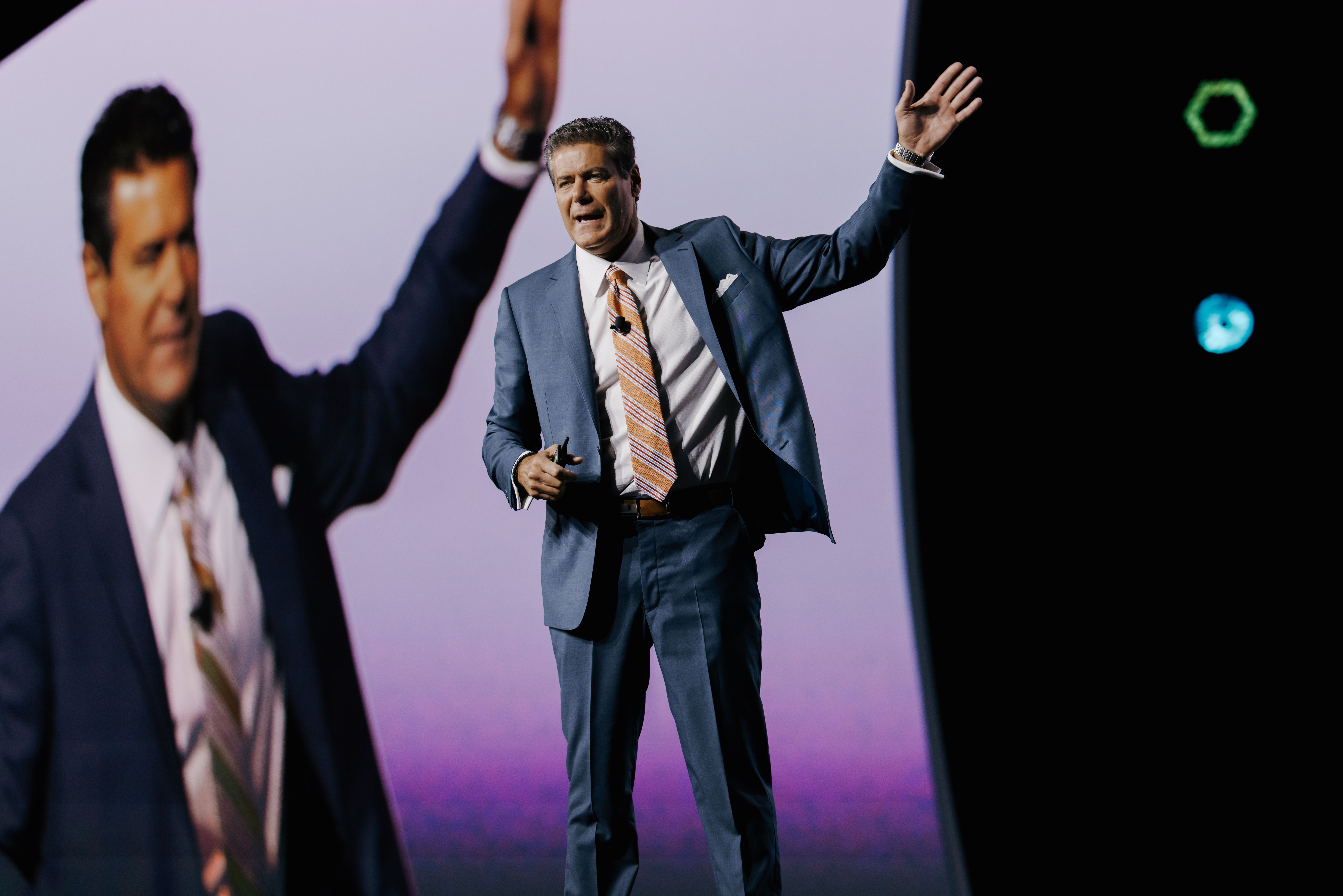 Philip Blancato speaking onstage with one arm raised and his image projected behind him