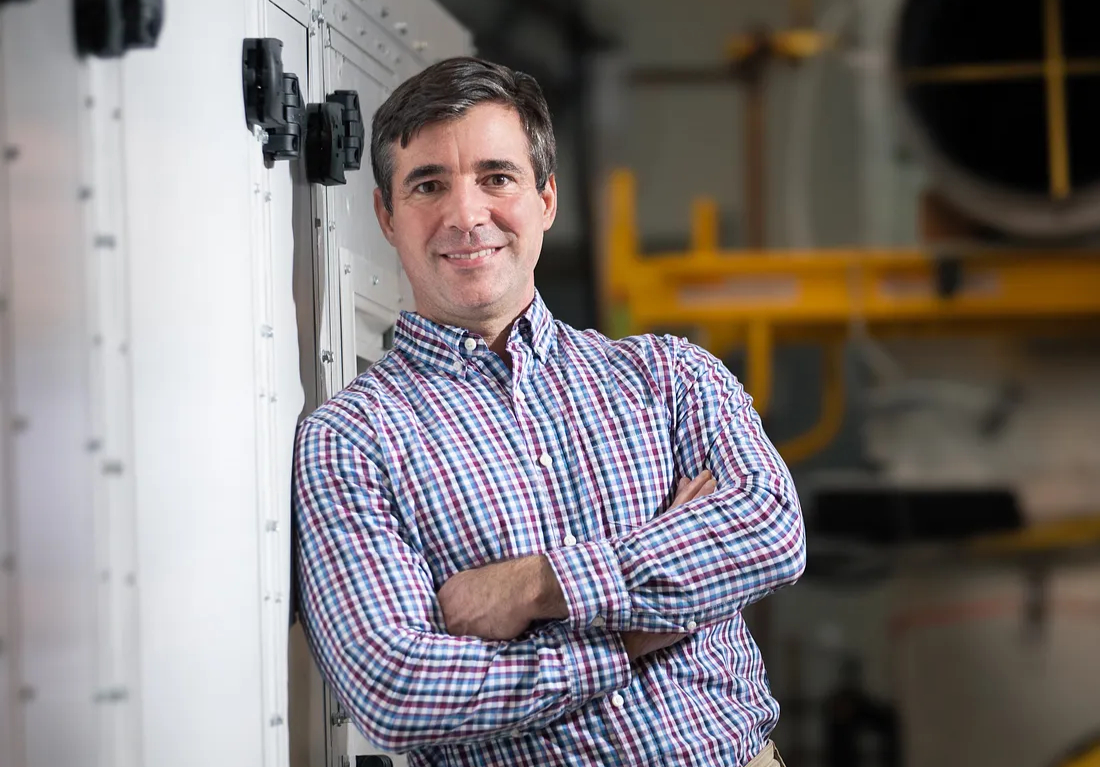 Philip Farese in a checked shirt, leaning beside a large HVAC unit in an industrial setting