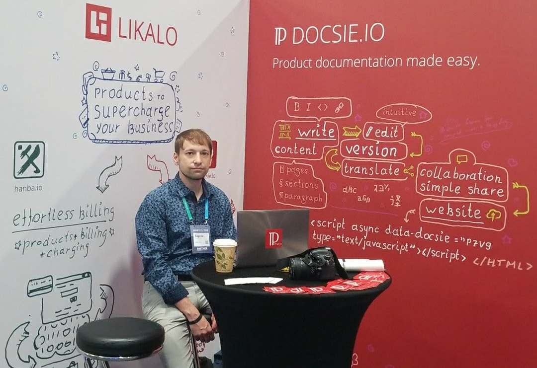 Philippe Trounev seated at an exhibition booth with Docsie and hanba.io displays, a laptop and a coffee cup