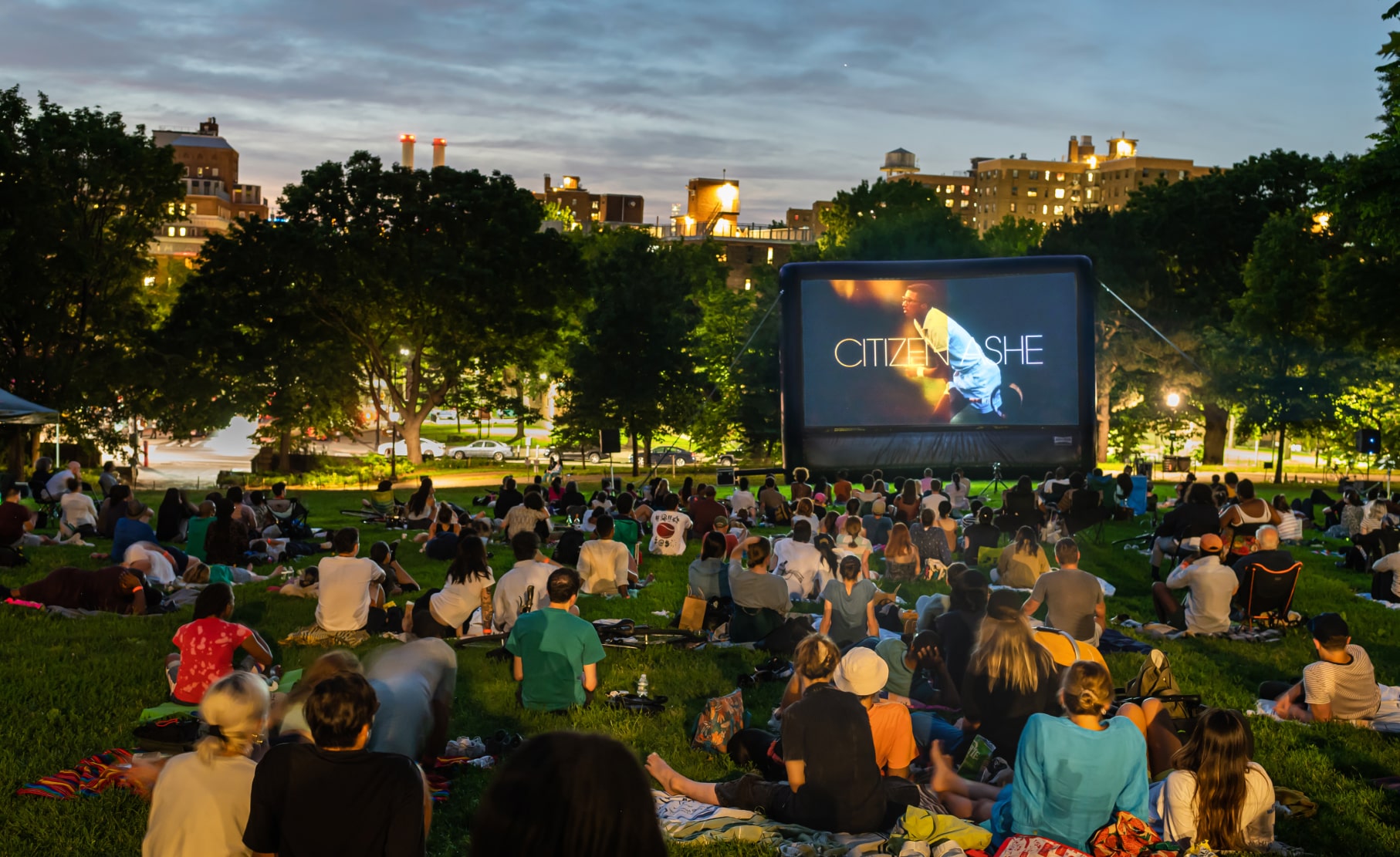 A large audience watches Citizen Ashe on an outdoor screen in a city park