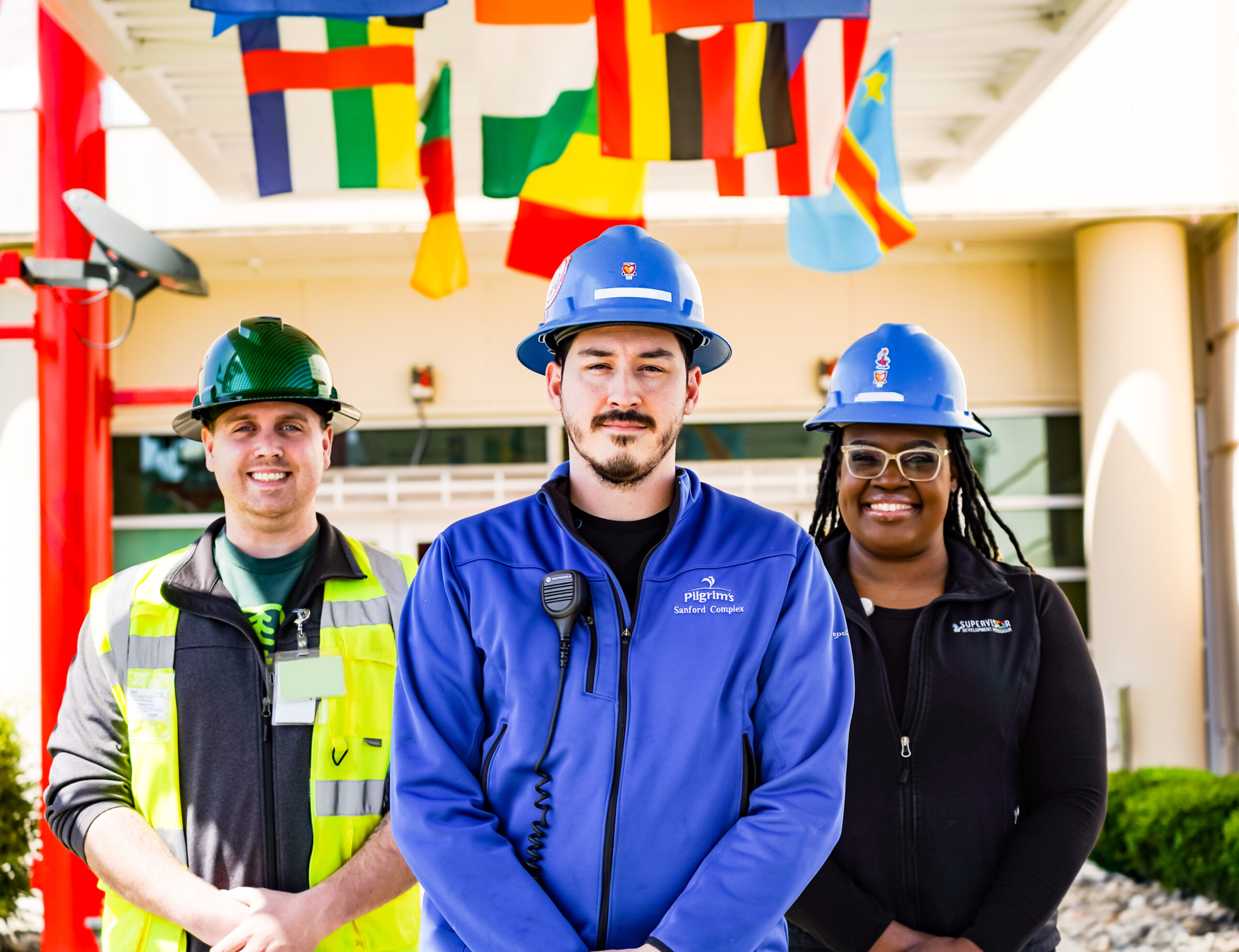 Three Pilgrim’s team members wearing hard hats, standing beneath international flags