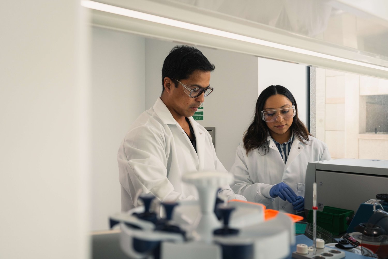 Two people in laboratory coats and protective glasses working beside laboratory equipment