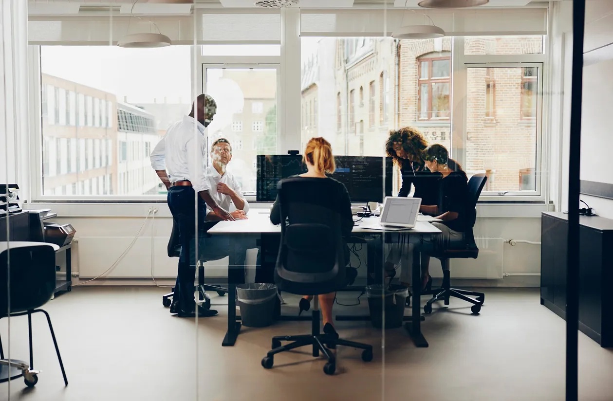 An illustrative office scene with colleagues gathered around desks behind a glass wall.