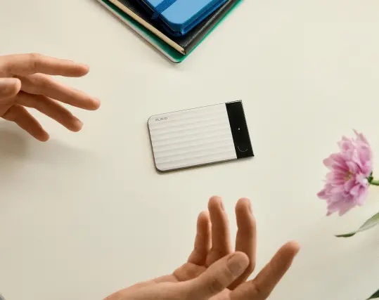 A silver Plaud Note Pro sits among notebooks and gesturing hands on a meeting table.